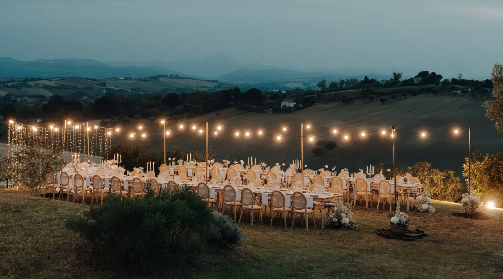Wedding dinner tables under string lights at dusk, overlooking the hills of Le Marche
